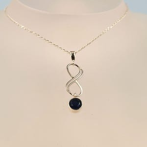 May include: A silver-toned necklace featuring a delicate chain and a vertical infinity symbol pendant. The pendant is accented with a single, round, dark blue faceted gemstone suspended from the bottom loop of the infinity design.