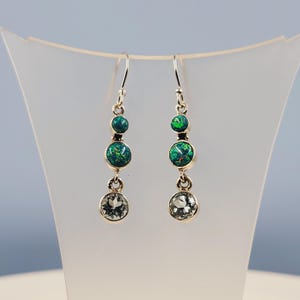May include: A pair of silver dangle earrings with three green opal stones and a single clear stone on each earring.