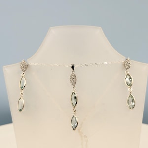 Op de afbeelding: Een zilveren ketting- en oorbellen set gepresenteerd op een witte mannequin. De set bevat een hanger en oorbellen met een marquise-vormig, lichtblauw edelsteenontwerp. De oorbellen en hanger zijn verbonden met een fijne zilveren ketting.
