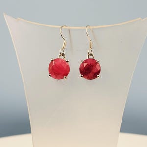 May include: A pair of dangle earrings featuring faceted, round, deep red gemstones. The gemstones are held in place by silver-colored prongs and suspended from silver-colored fish hook ear wires. The earrings are displayed on a white, curved stand.