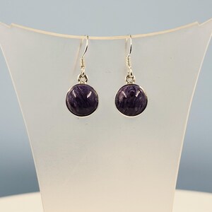 Peut inclure: Une paire de boucles d'oreilles pendantes avec des pierres précieuses charoïte violettes, polies et rondes, serties dans des montures en métal argenté. Les pierres présentent des motifs tourbillonnants blancs et violet foncé.