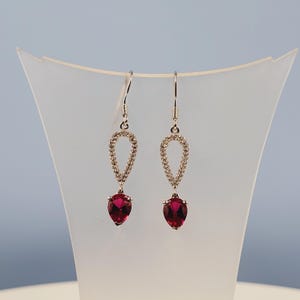 May include: A pair of silver-toned drop earrings. Each earring features a teardrop-shaped, deep red gemstone suspended below a loop of small, clear stones. The earrings hang from a simple hook.