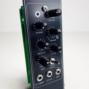 Peut inclure: Un module de synthétiseur modulaire noir étiqueté SYNDRUM et CORON DS-8 CLONE. La façade comporte six boutons rotatifs noirs pour decay, sweep, LFO speed, LFO amount, V.C.O. et noise pan, un interrupteur à bascule en métal et trois prises jack.