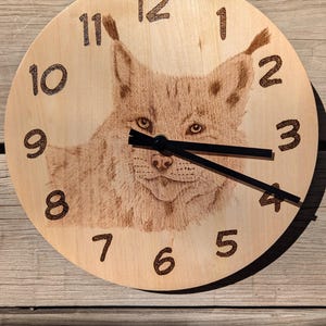 May include: Wooden wall clock featuring a detailed engraving of a lynx. The clock face is light brown with dark brown numerals and black clock hands. The lynx image is centered, with its face and ears prominently displayed.