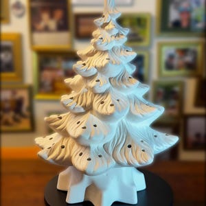 May include: A white ceramic Christmas tree with a star-shaped base. The tree has detailed, layered branches with small holes for lights. The tree is displayed on a black circular stand, perfect for holiday decor.