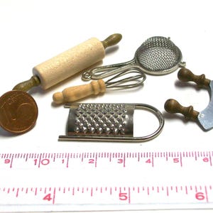 Pode incluir: Uma coleção de utensílios de cozinha em miniatura, incluindo um rolo de massa de madeira, uma peneira de metal, um batedor, um ralador de metal e uma lâmina curva com cabos de madeira, ao lado de uma moeda de 10 cêntimos e uma régua.