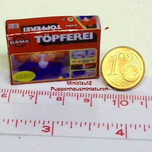 Puede incluir: Una caja roja con las palabras "TÖPFEREI" en letras blancas. La caja contiene un kit de alfarería para hacer cerámica en miniatura. Una moneda de 1 céntimo de euro está al lado de la caja para la escala.