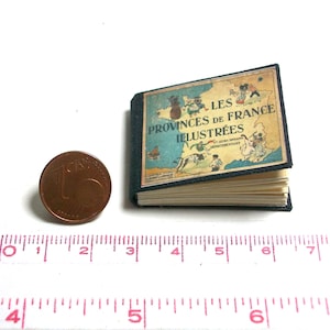 Könnte beinhalten: Ein kleines, blaues, gebundenes Buch mit dem Titel "Les Provinces de France Illustrées" auf dem Einband. Das Buch ist geschlossen und liegt auf einer weißen Oberfläche neben einer kupferfarbenen Münze.