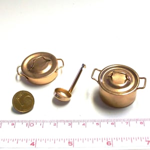 Può includere: Set di pentole in miniatura color rame, composto da due pentole con coperchio e un mestolo. Una moneta è accanto per il confronto delle dimensioni. Il set è posto su una superficie bianca.