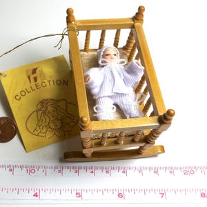 Könnte beinhalten: Eine Miniatur-Schaukelwiege aus Holz mit einer kleinen Puppe in weißer Kleidung. Die Puppe trägt einen Hut und liegt auf einem beigen Kissen. Ein Etikett mit dem Wort "COLLECTION" ist an der Wiege befestigt.