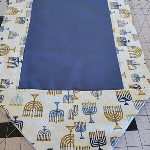 Puede incluir: Un camino de mesa de tela azul con fondo blanco y un patrón de menorás azules y dorados. El camino de mesa tiene un borde de tela azul.