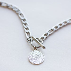 May include: A silver necklace with a chain link design and a circular pendant. The pendant has an engraved pattern and is attached to the chain with a toggle clasp. The necklace is shown on a white surface.
