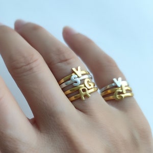 Peut inclure: Une main portant un ensemble de quatre bagues initiales. Les bagues sont empilées sur l'annulaire. Les deux bagues du haut sont en argent et portent les lettres "V" et "S". Les deux bagues du bas sont en or et portent les lettres "R" et "G". Les bagues sont étiquetées "Bagues initiales en or et en argent".