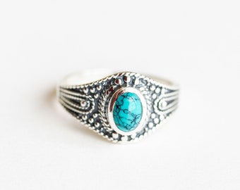 Peut inclure: Bague en argent ornée d'une pierre ovale turquoise. La bague présente un motif ornemental et détaillé autour de la pierre et sur le corps. La pierre turquoise est d'un bleu-vert vif. La bague est posée sur un fond blanc.