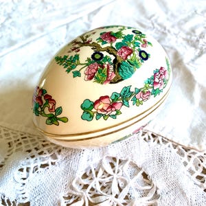 May include: Egg-shaped decorative box in cream with a floral design. The box has pink, green, and blue flowers with gold trim. The floral pattern is set against a cream background, creating a vintage aesthetic.