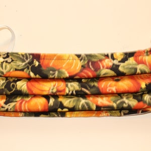 May include: A black fabric face mask with a pumpkin pattern. The mask has white elastic ear loops.
