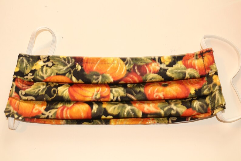 May include: A black fabric face mask with a pumpkin pattern. The mask has white elastic ear loops.