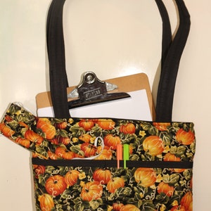 May include: A black tote bag with a pumpkin print and black straps. The bag has multiple pockets and is filled with a clipboard and pens.
