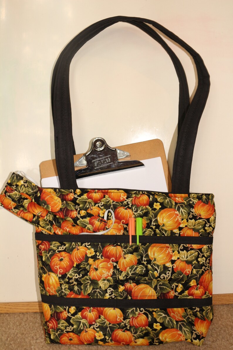 May include: A black tote bag with a pumpkin print and black straps. The bag has multiple pockets and is filled with a clipboard and pens.