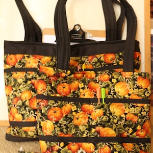 May include: Two black tote bags with pumpkin and vine patterned fabric. The bags have multiple pockets and black straps. A matching fabric face mask is in the foreground.