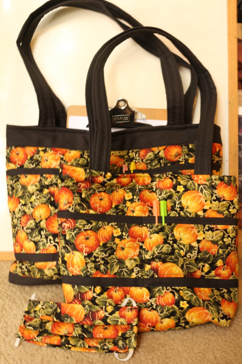 May include: Two black tote bags with pumpkin and vine patterned fabric. The bags have multiple pockets and black straps. A matching fabric face mask is in the foreground.
