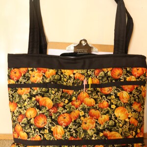 May include: A black tote bag with a pumpkin print and black straps. The bag has multiple pockets and a black zipper closure. The bag is sitting on a brown carpet.