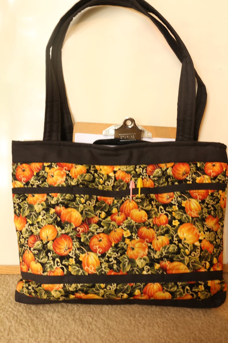 May include: A black tote bag with a pumpkin print and black straps. The bag has multiple pockets and a black zipper closure. The bag is sitting on a brown carpet.