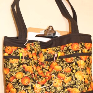 May include: Black tote bag with a pumpkin and vine pattern. The bag has multiple pockets and a black strap. A clipboard is inside the bag.
