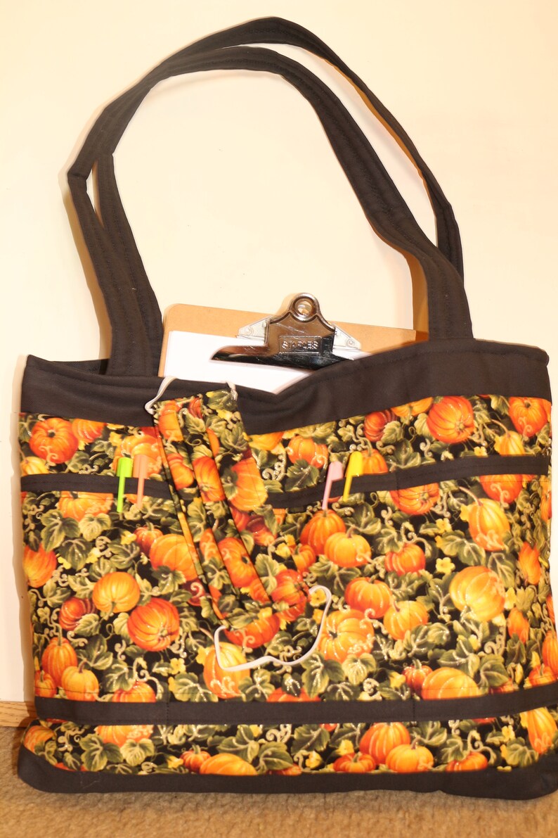 May include: Black tote bag with a pumpkin and vine pattern. The bag has multiple pockets and a black strap. A clipboard is inside the bag.
