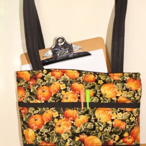 May include: A black tote bag with a pumpkin print and black trim. The bag has three pockets on the front and a clipboard with a white sheet of paper inside.