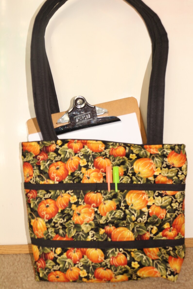 May include: A black tote bag with a pumpkin print and black trim. The bag has three pockets on the front and a clipboard with a white sheet of paper inside.