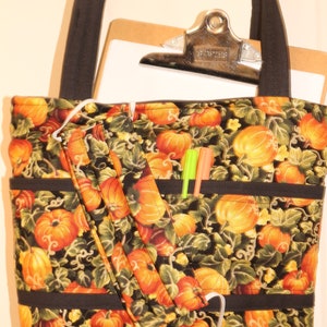 May include: A black tote bag with a pumpkin print. The bag has a black strap and a zippered pocket. There are two pens in the pocket. The bag is made of fabric and has a white lining.