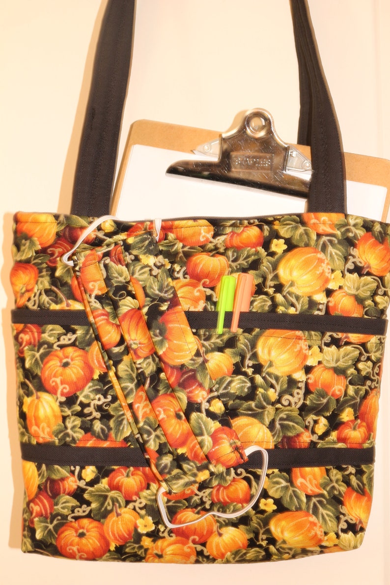 May include: A black tote bag with a pumpkin print. The bag has a black strap and a zippered pocket. There are two pens in the pocket. The bag is made of fabric and has a white lining.