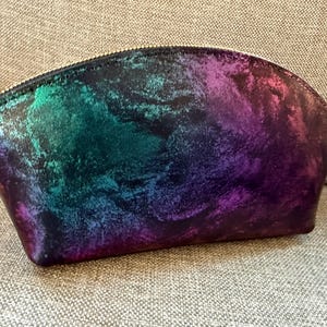May include: A cosmetic bag with a colorful, abstract design. The bag features a zipper closure and a vibrant mix of colors, including shades of purple, green, and blue. The bag is made of a shiny material.