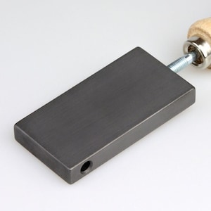 May include: A grey rectangular metal block with a hole on one side and a wooden handle attached to the top with a screw.