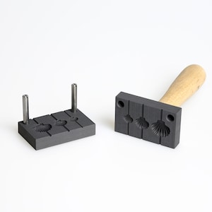 May include: A two-piece mold for making seashell-shaped jewelry. The mold is made of black graphite and has a wooden handle. The mold is open and ready to be filled with molten metal.