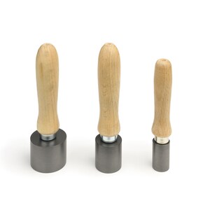May include: Three metal tools with wooden handles of varying sizes. The handles are light brown, and the cylindrical metal bases are dark grey. The tools are arranged vertically against a white background.