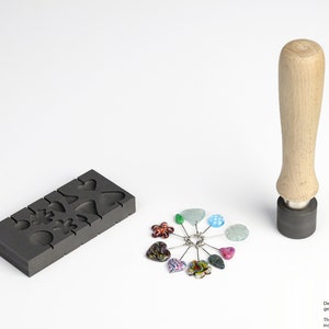 May include: A black graphite mould with various heart, flower, and leaf shapes. A wooden handle with a black base and a set of colourful glass beads are also shown. The text "Die hier abgebildeten Glasperlen gehören nicht zum Lieferumfang The glassbeads illustrated here are not induded in the scope of delivery!" is printed below the image.