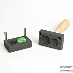 Puede incluir: Un molde negro con dos cavidades en forma de trébol, una con una cuenta de vidrio verde en su interior. El molde está hecho de grafito y tiene un mango de madera. El texto "¡La cuenta de vidrio ilustrada aquí no está incluida en el alcance de la entrega!" está escrito debajo del molde.