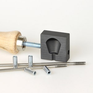 Può includere: Una collezione di utensili e componenti hardware in metallo. L'immagine presenta uno strumento con manico in legno e asta filettata, uno stampo in metallo grigio scuro e diversi piccoli cilindri e aste metalliche. Gli oggetti sono disposti su una superficie bianca.