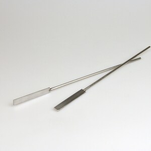 May include: Two thin, silver metal spatulas with flat, rectangular blades. The spatulas are crossed over each other.