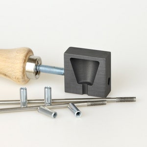 Peut inclure: Un moule en métal avec une poignée en bois, utilisé pour fabriquer des inserts métalliques. Le moule est gris et de forme carrée. La poignée est brune et a une tige filetée attachée à elle. Il y a aussi plusieurs inserts métalliques dans l'image.