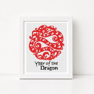 May include: A framed cross-stitch embroidery featuring a circular red dragon motif with intricate swirling patterns. Below the dragon, the text Year of the Dragon is stitched in black thread. The entire design is set against a white fabric background.
