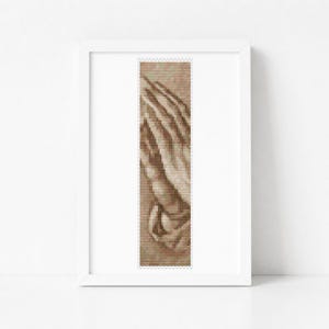 May include: A framed vertical cross-stitch pattern featuring a sepia-toned image of two hands pressed together in a prayer position. The design is composed of small, uniform stitches in shades of brown and beige, centered within a white frame.