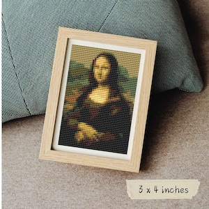 May include: A framed cross-stitch of the Mona Lisa, in a light wood frame. The artwork is a pixelated version of the famous painting. The frame measures 3 x 4 inches.
