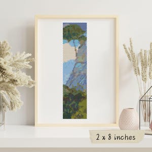 Op de afbeelding: Een kruissteekpatroon van een landschap met een blauwe lucht, groene bomen en een berg op de achtergrond. Het patroon is ingelijst in een licht houten lijst en meet 5,1 x 20,3 cm.