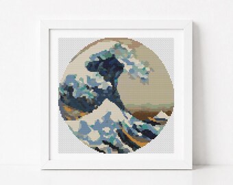 May include: A square white frame containing a circular cross-stitch embroidery piece. The design depicts a stylised ocean wave in shades of dark blue, light blue, and white, set against a beige background, mimicking a classic Japanese woodblock print.