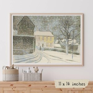 May include: Framed winter scene print depicting a snowy village with buildings and a road. The artwork is in a light wood frame and measures 28 x 36 cm. The print shows a winter landscape with falling snow.