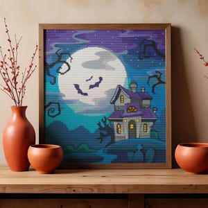 May include: A square framed cross-stitch embroidery depicting a haunted house under a large full moon in a purple and blue night sky. The scene features bare, twisted tree branches, a small graveyard, and a house with glowing yellow windows.
