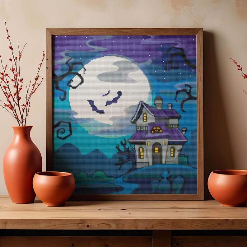 May include: A square framed cross-stitch embroidery depicting a haunted house under a large full moon in a purple and blue night sky. The scene features bare, twisted tree branches, a small graveyard, and a house with glowing yellow windows.